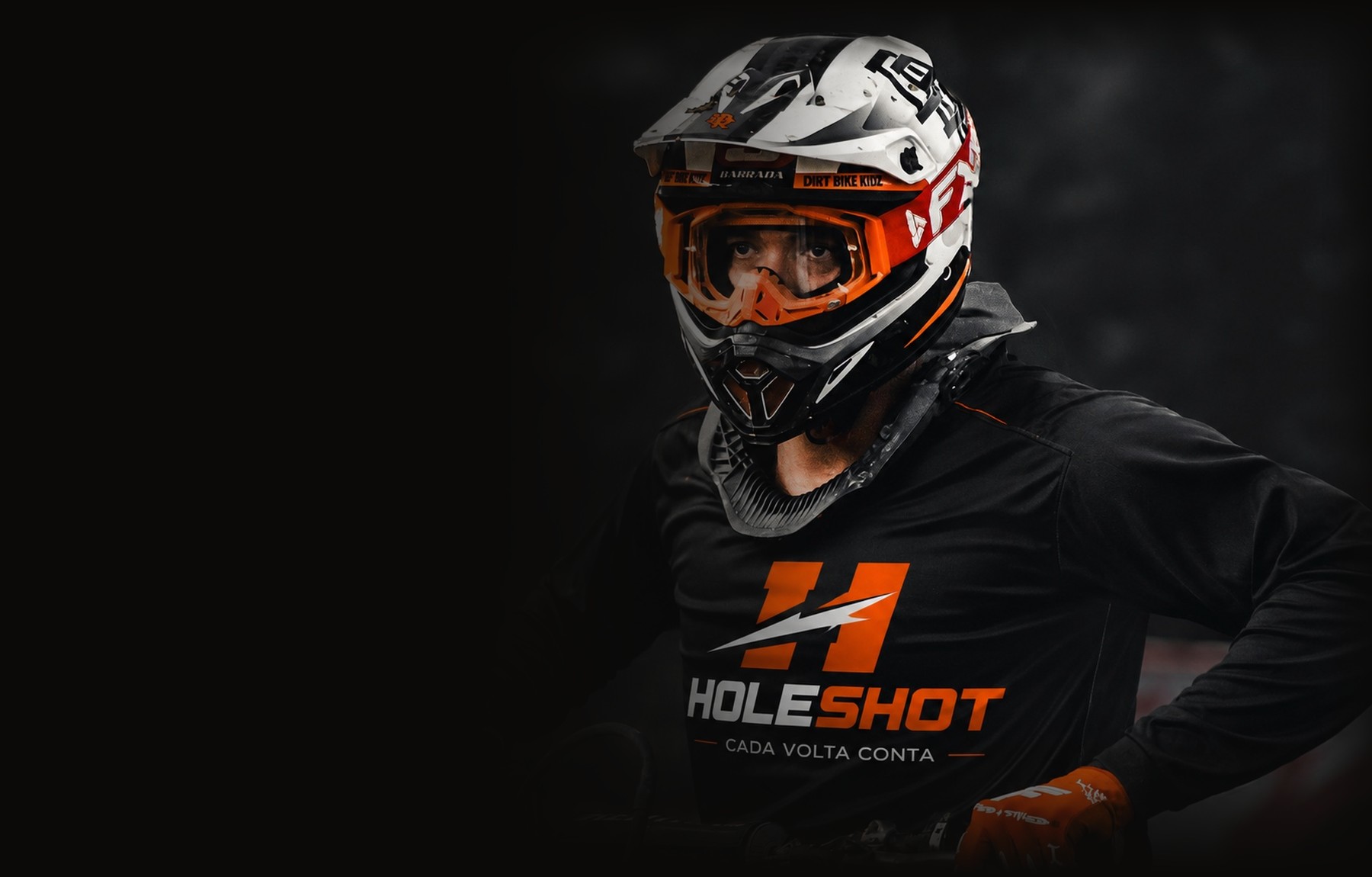 Piloto com camisa HOLESHOT, capacete e óculos, apoiado na moto antes de entrar na pista